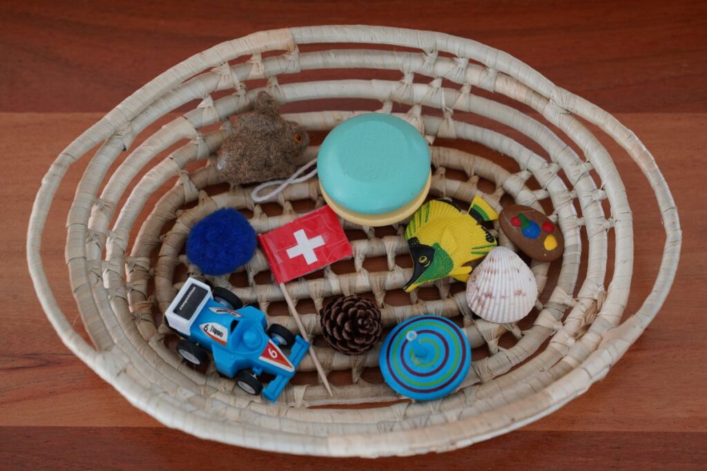 Basket of small objects for Montessori Sound Games, including a toy car, shell, pinecone, flag, and other items to support phonetic awareness and early language development.