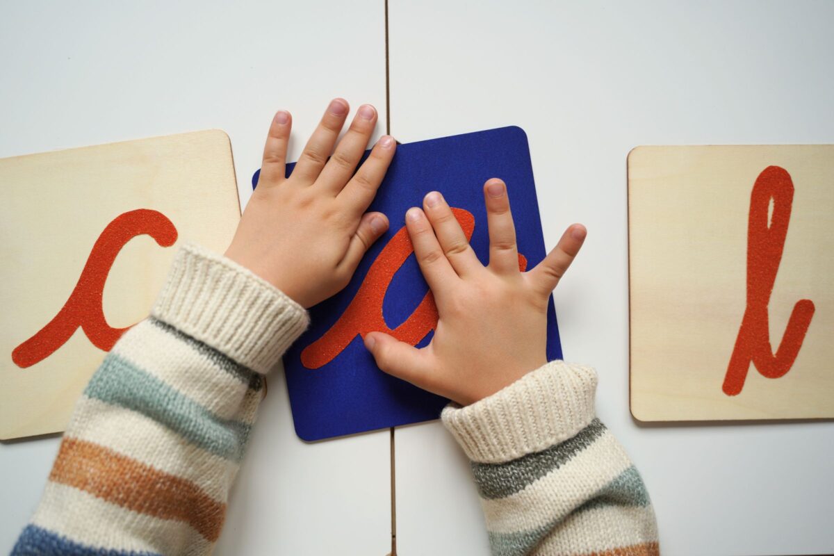 Child practicing Montessori sandpaper letters activity, tracing an orange cursive letter on blue wood