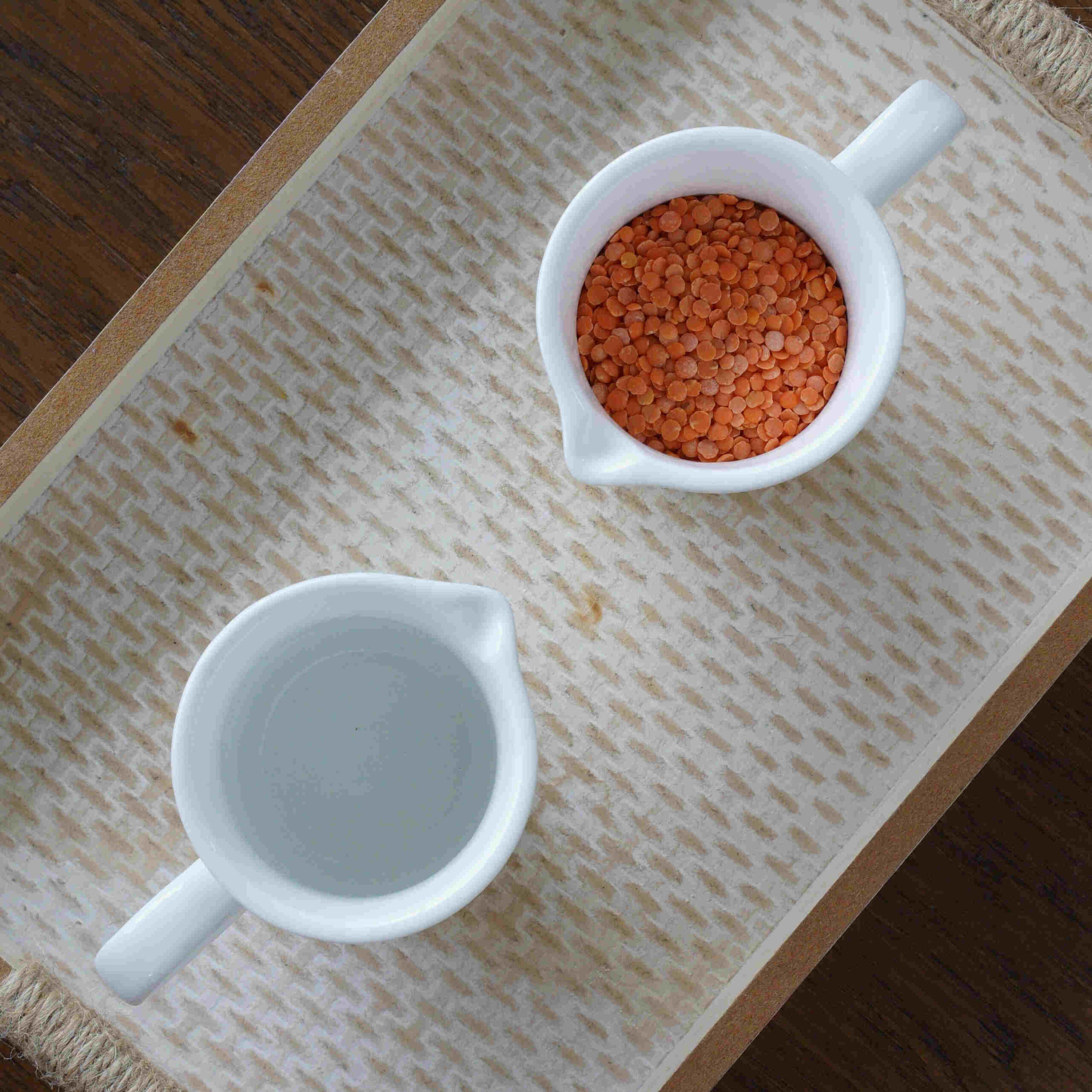 Two small pitchers sitting on a tray ready to be used for a Montessori activity. One pitcher is filled with red lentils and the other pitcher is empty.