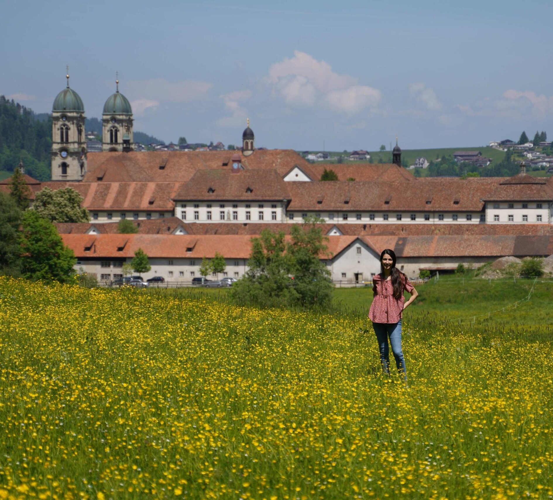 A young woman standing in a yellow dandelion covered field in front of the Einsiedeln monastery.