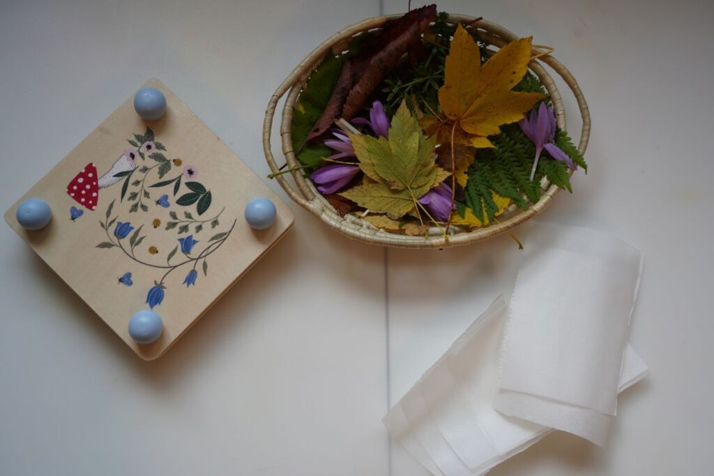 Materials for a Montessori-inspired flower pressing activity, including a wooden flower press, basket of leaves and flowers, and pieces of parchment paper.