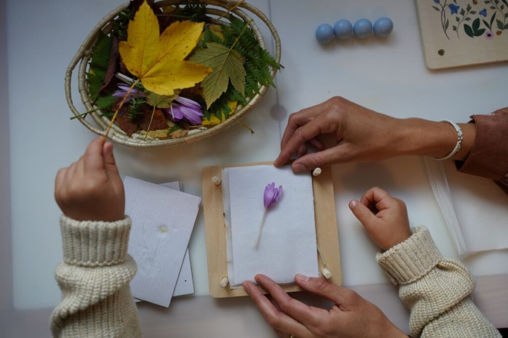 Adult and child pressing a purple flower between sheets of paper in a wooden flower press, with a basket of colorful autumn leaves and flowers nearby.
