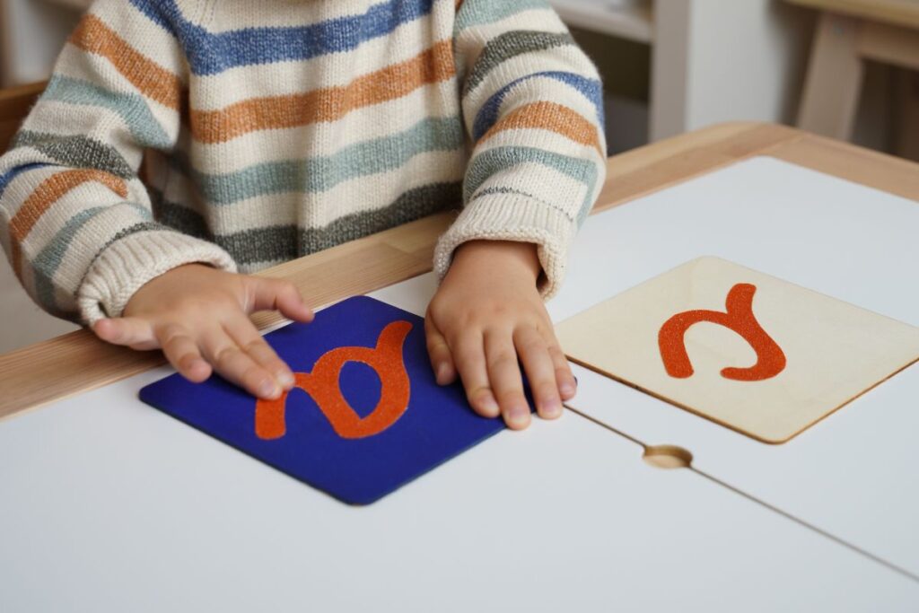 A young child traces sandpaper letters on Montessori cards, practicing letter sounds and tactile learning at a table.