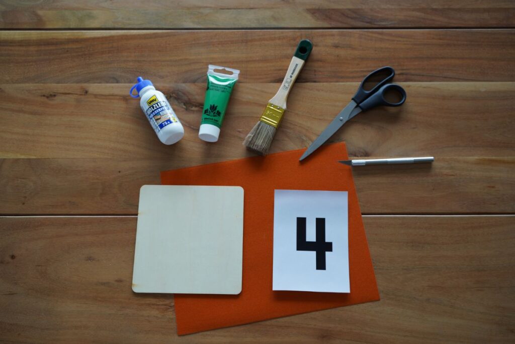 Materials for a DIY Montessori Sandpaper Numbers activity, including glue, green paint, brush, scissors, utility knife, wooden board, sandpaper, and printed number 4 template.