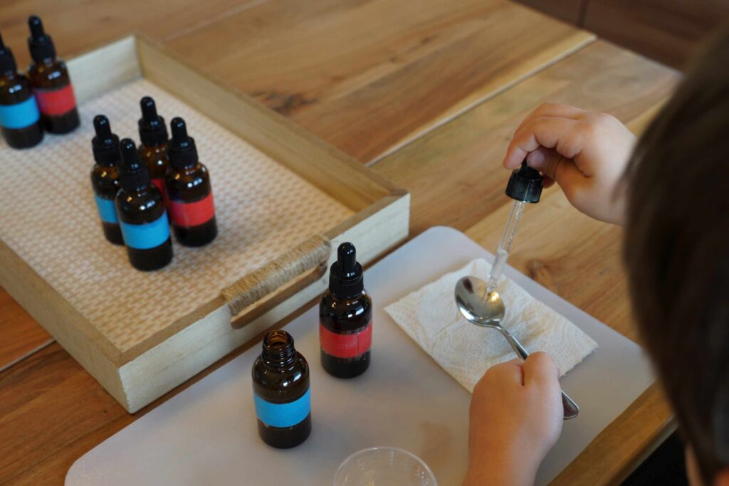 Child using a dropper to place liquid onto a spoon during a Montessori tasting bottles activity, with color-coded bottles arranged on a wooden tray.