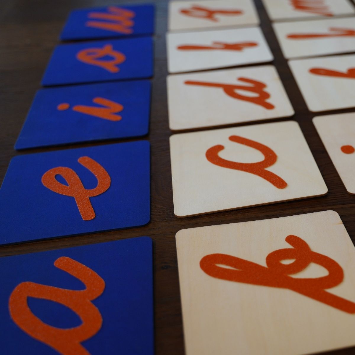 Close-up of Montessori sandpaper letters showing lowercase cursive letters in orange textured material on wooden boards, with vowels on blue backgrounds and consonants on natural wood, arranged neatly on a dark wooden surface.
