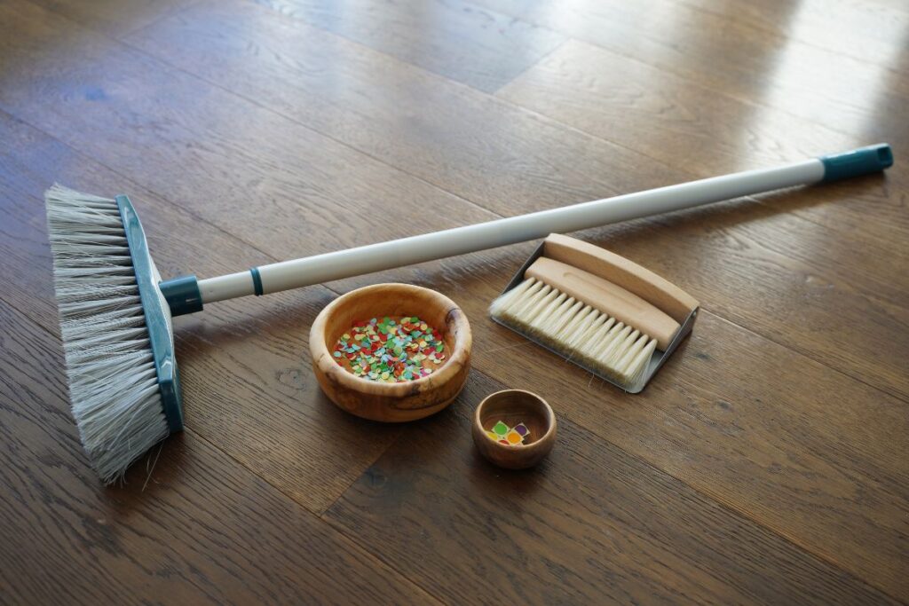 Montessori sweeping floor activity with child-sized broom, dustpan brush, and wooden bowls for practical life cleaning exercise and fine motor skill development.