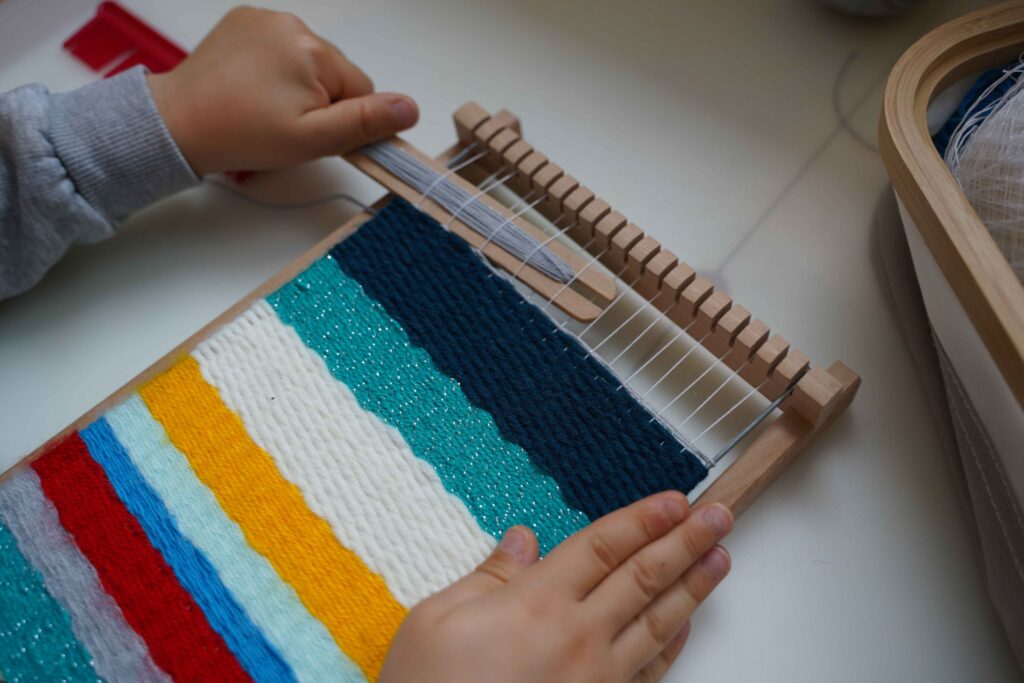 Child weaving colorful yarn on a small wooden loom, creating bright textured stripes as part of a Montessori-inspired hands-on fine motor and practical life activity.