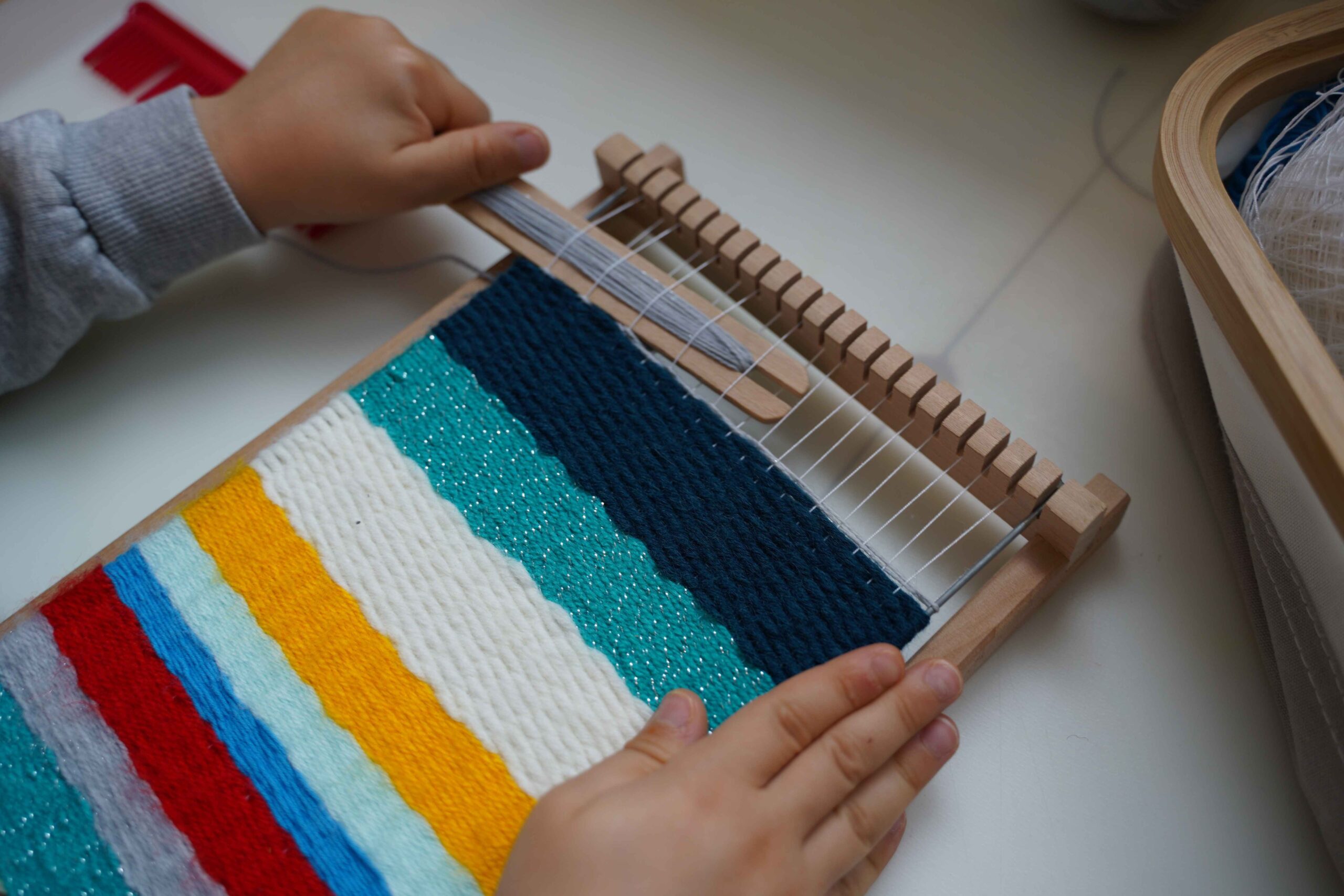 Child weaving colorful yarn on a small wooden loom, creating bright textured stripes as part of a Montessori-inspired hands-on fine motor and practical life activity.
