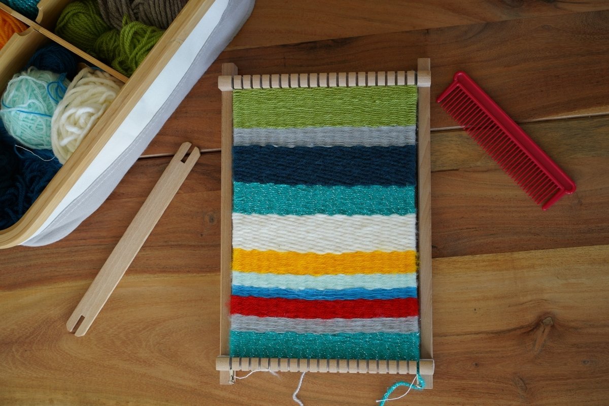 Colorful finished weaving on a wooden loom with yarn in shades of red, yellow, blue, teal, gray, and green, surrounded by weaving tools and yarn basket as part of a Montessori-inspired textile activity.