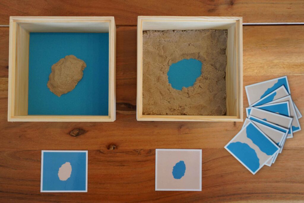 Montessori land and water forms activity showing wooden boxes with sand and blue paper illustrating an island and a lake, alongside matching geography cards.