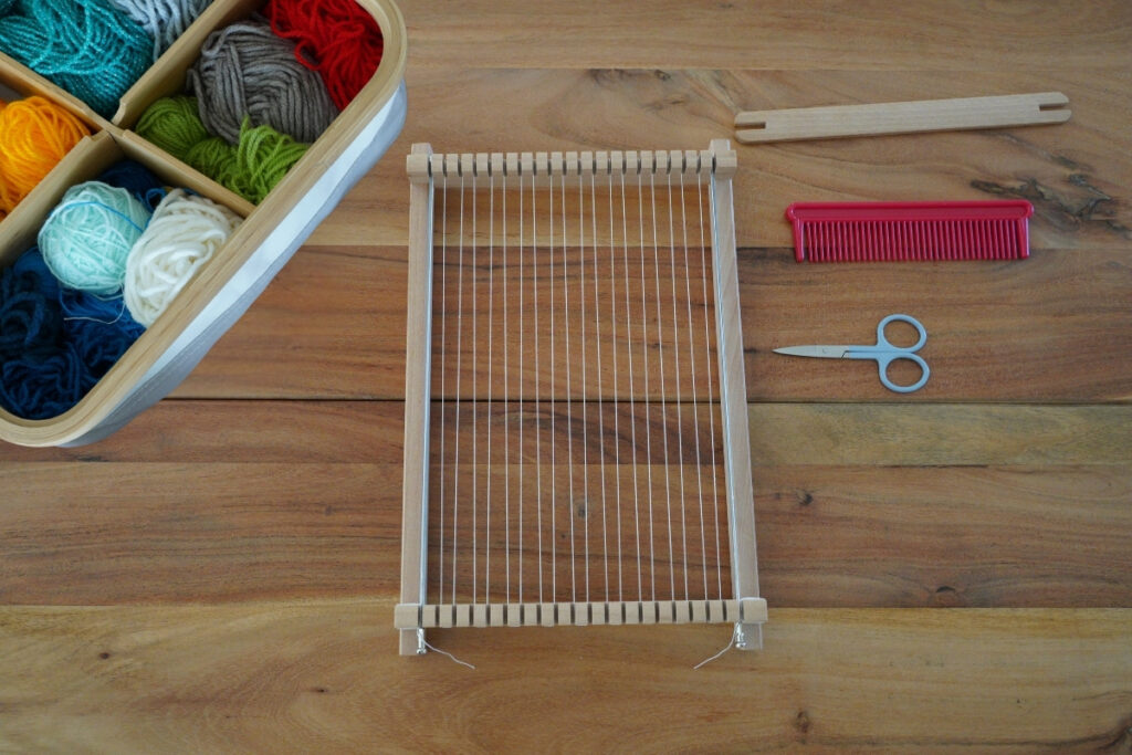 Montessori weaving setup with a wooden loom strung with warp threads, colorful yarn balls in a basket, scissors, a red comb, and a wooden shuttle arranged neatly on a wooden table ready for a hands-on crafting activity.