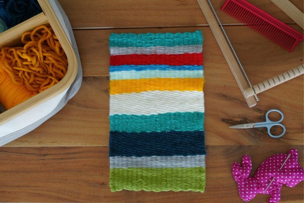 Completed colorful woven fabric piece made from a Montessori weaving activity, surrounded by yarn, scissors, a loom frame, and tools on a wooden table, showcasing a hands-on crafting project for children.