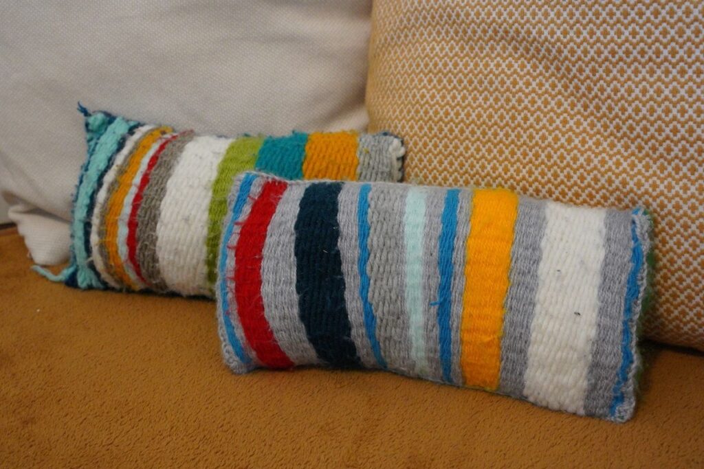 Two handmade pillows covered with colorful woven fabric created from a Montessori weaving project, featuring bright yarn stripes in red, blue, yellow, green, and white, displayed on a cozy rug.