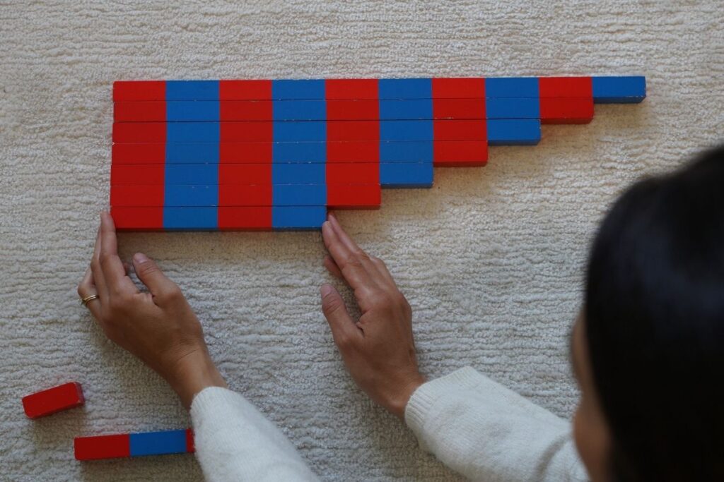 Woman arranging Montessori number rods in order on a light carpet to explore counting and quantity.