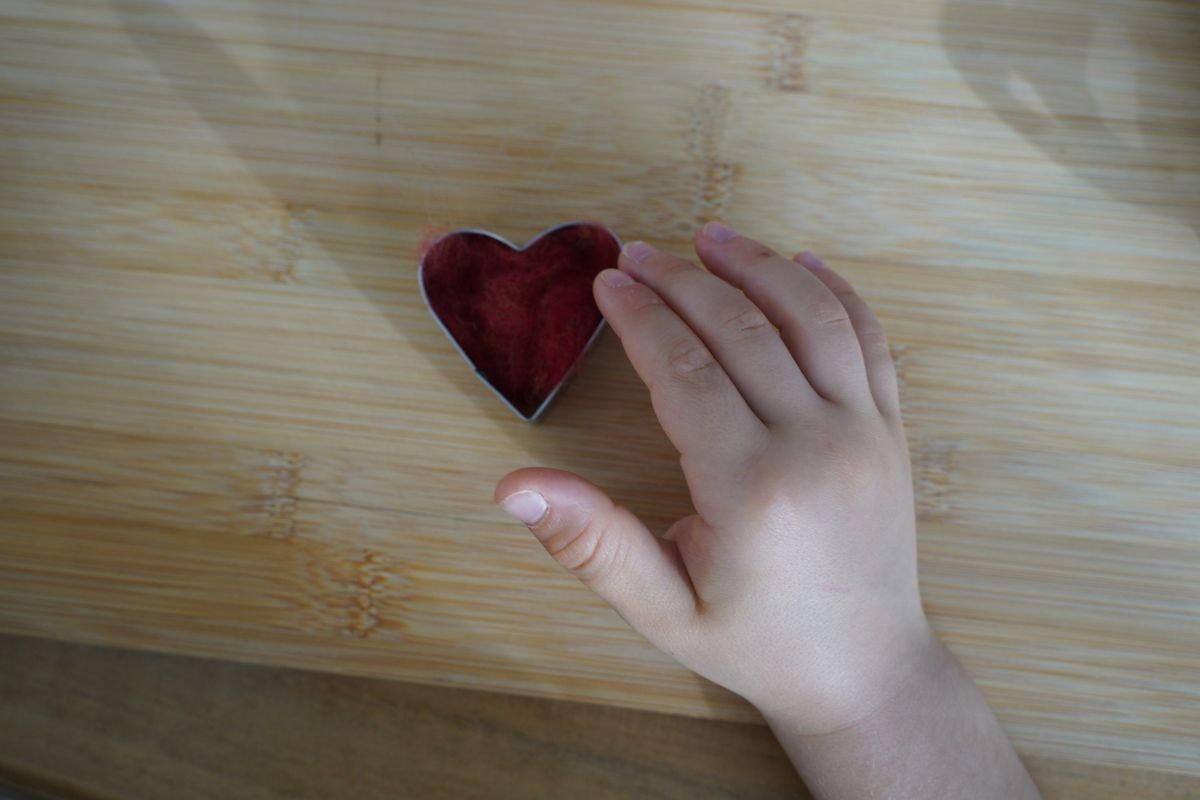 Child’s hand pressing wool roving into a heart-shaped cookie cutter during a Montessori wet felting activity for fine motor development.