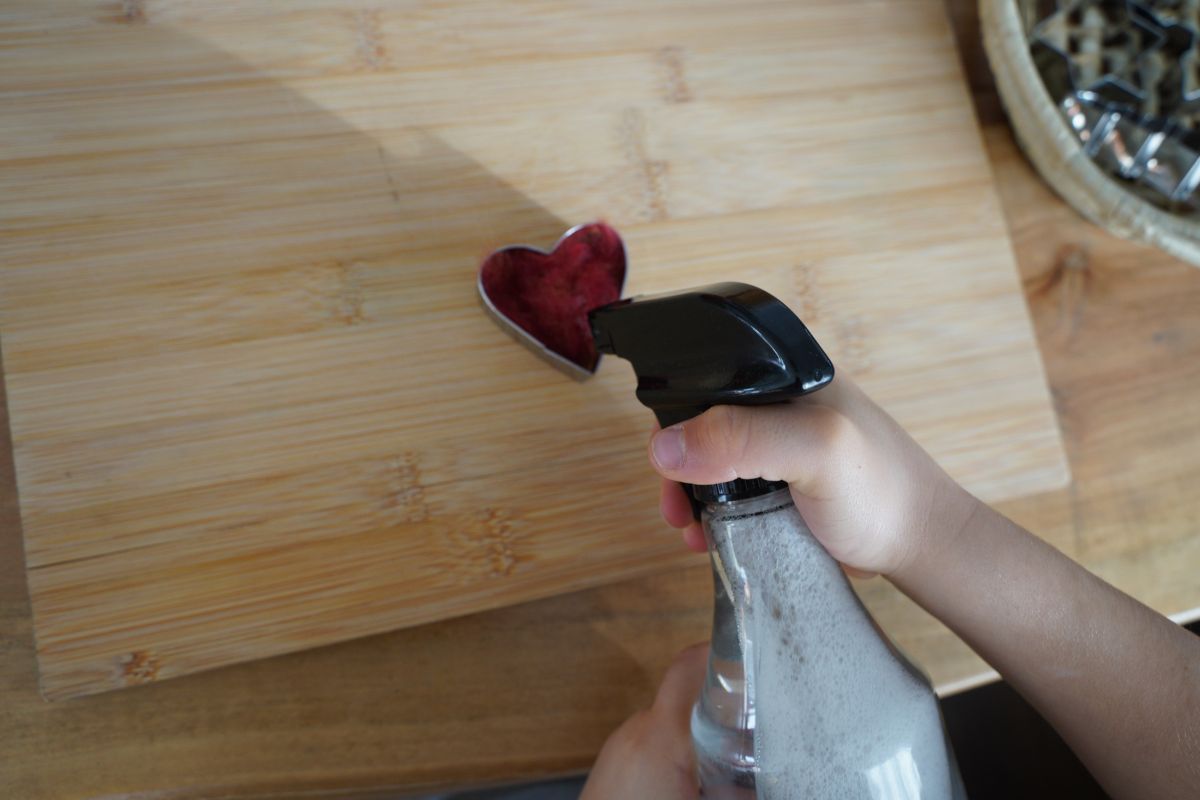 Child using a spray bottle to wet wool inside a heart-shaped cookie cutter during a Montessori wet felting craft.