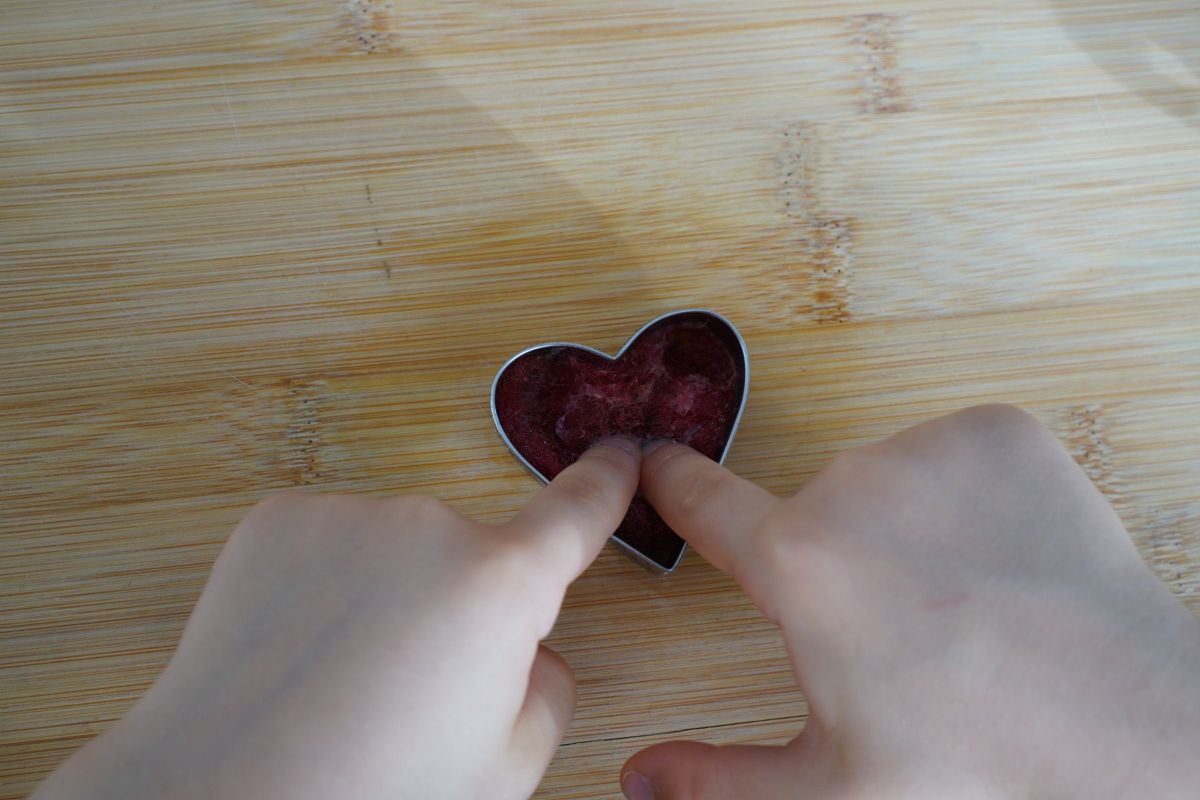 Child shaping and smoothing wool inside a heart cookie cutter as part of a Montessori wet felting craft.