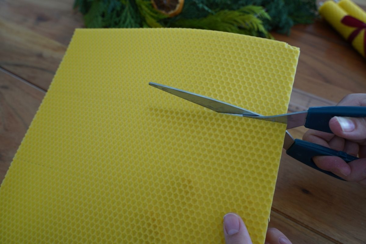 Cutting a beeswax sheet with scissors on a wooden table, preparing pieces for a Montessori candle-making practical life activity that develops fine motor control and hands-on learning with natural materials.