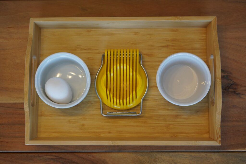 Montessori practical life egg slicing tray with a hard-boiled egg, egg slicer, and bowls for preparing food independently.