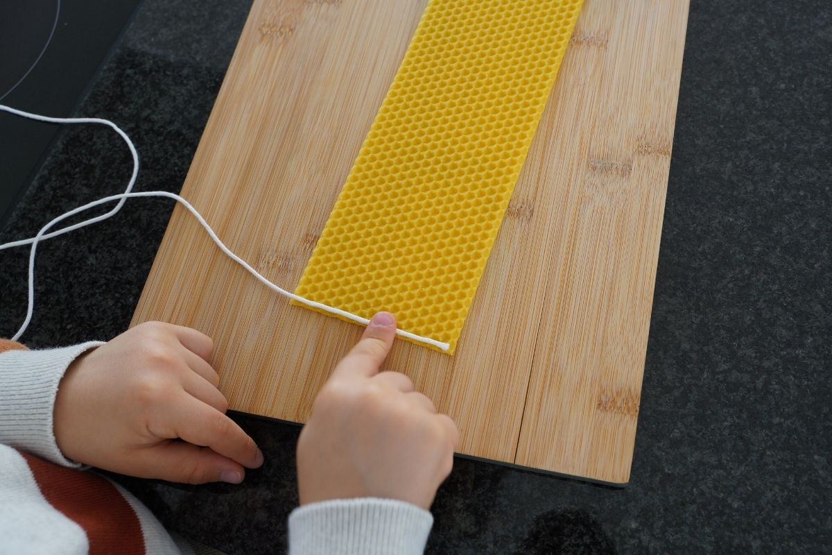 Child pressing a cotton wick into a beeswax sheet on a wooden board, beginning a Montessori candle-making practical life activity that supports fine motor skills and hands-on learning with natural materials.