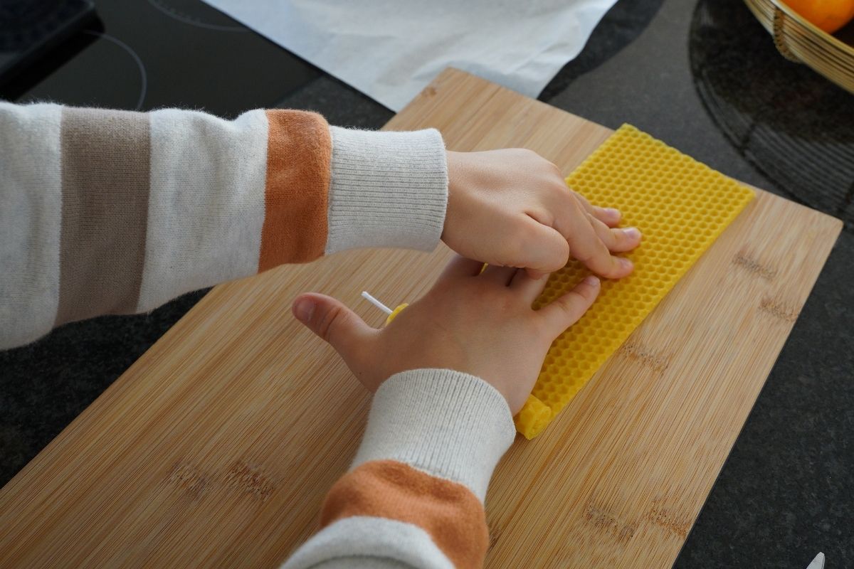 Child rolling a beeswax sheet around a cotton wick on a wooden board, completing a Montessori candle-making practical life activity that builds fine motor coordination and hands-on learning with natural materials.