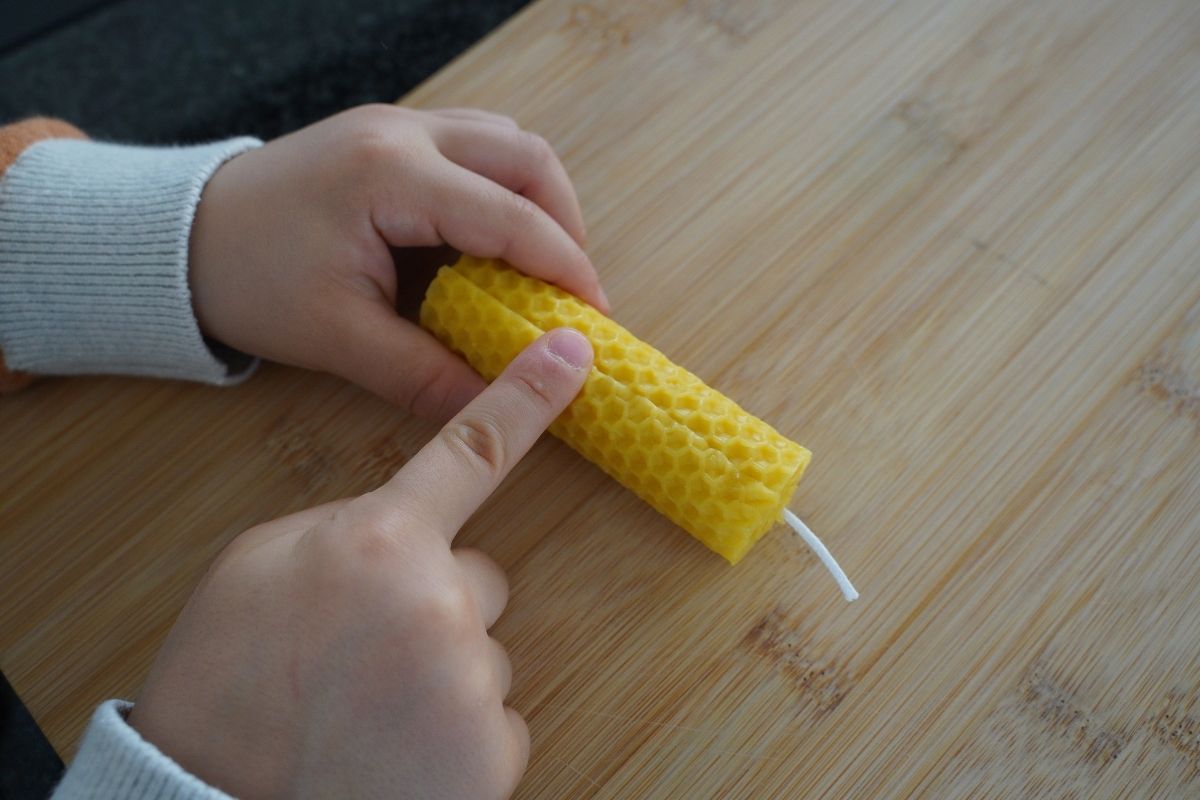 Child pressing and sealing the edge of a rolled beeswax candle with a finger on a wooden board, completing a Montessori candle-making practical life activity that strengthens fine motor skills and hands-on learning with natural materials.