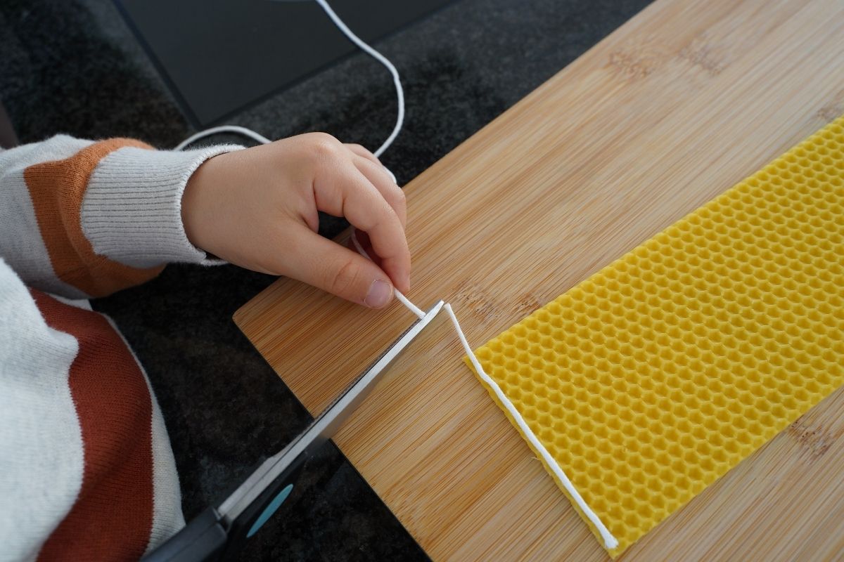 Child cutting a cotton candle wick with scissors next to a beeswax sheet on a wooden board, part of a Montessori candle-making practical life activity that develops fine motor skills and safe tool use through hands-on learning.
