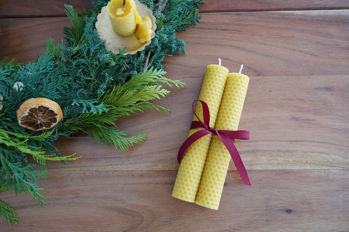 Handmade rolled beeswax candles tied with a red ribbon beside a natural evergreen Christmas wreath, showcasing a simple, hands-on Montessori-inspired craft using natural materials.