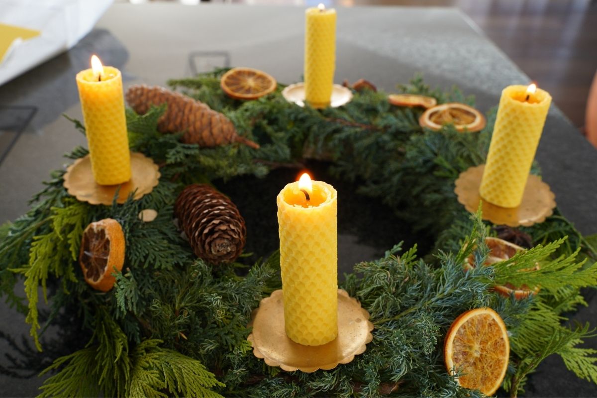 Four handmade rolled beeswax candles lit in a natural evergreen Christmas wreath with pinecones and dried oranges, showcasing a Montessori practical life activity using natural materials and hands-on candle making for children.