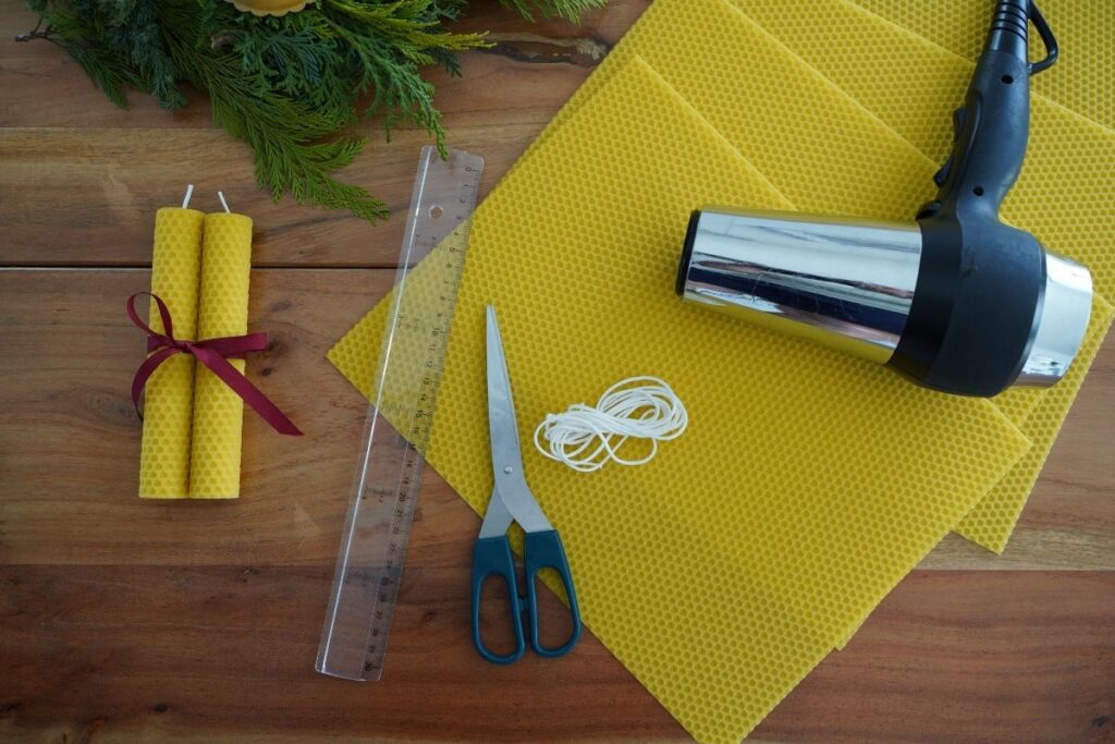 Materials for a Montessori beeswax candle-making activity, including yellow beeswax sheets, cotton wicks, scissors, ruler, hair dryer for warming wax, and finished rolled candles, arranged on a wooden table for a hands-on practical life activity.
