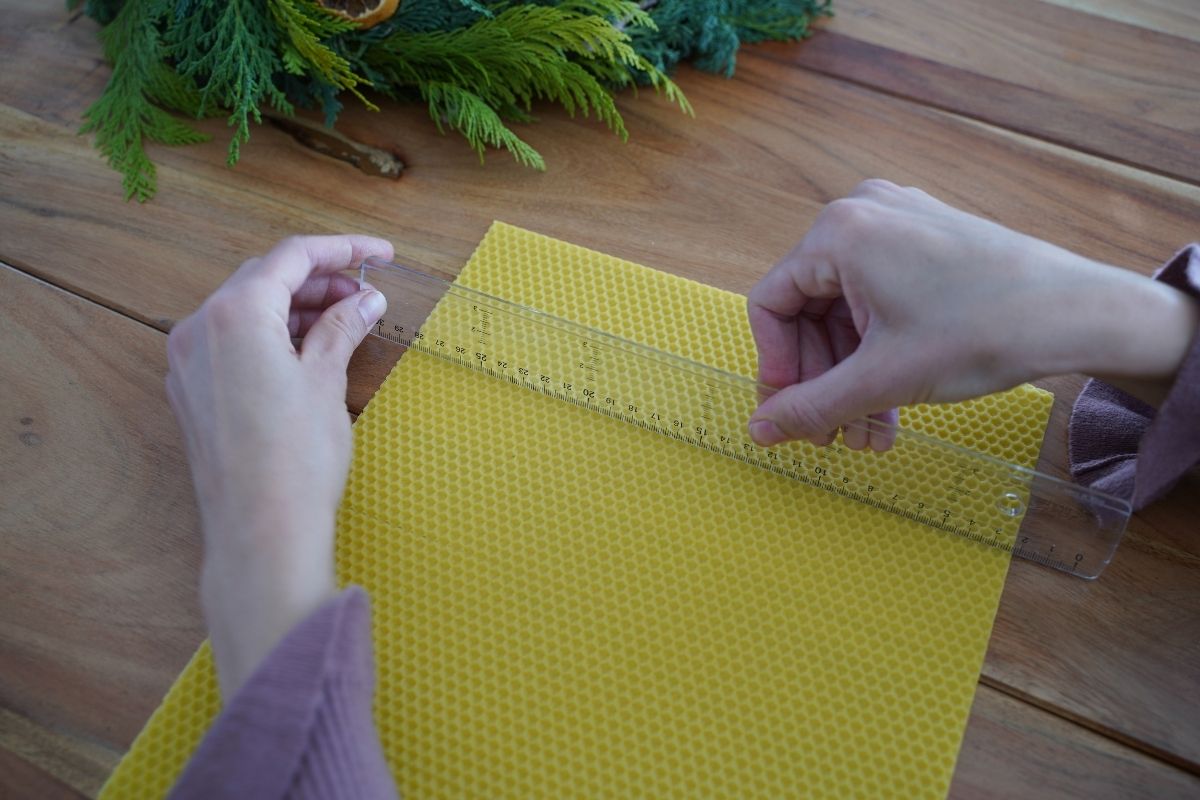 Measuring a beeswax sheet with a ruler on a wooden table, preparing materials for a Montessori candle-making practical life activity that supports math skills, precision, and hands-on learning with natural materials.