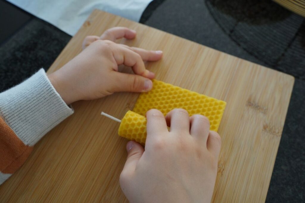 Child rolling a beeswax candle by hand on a wooden board, practicing a Montessori practical life activity that builds fine motor skills through hands-on candle making with natural materials.