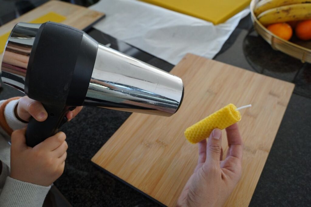Child gently warming a rolled beeswax candle with a hair dryer while an adult holds the candle, sealing the end of the candle during a Montessori practical life activity that teaches safe tool use and hands-on candle making with natural materials.
